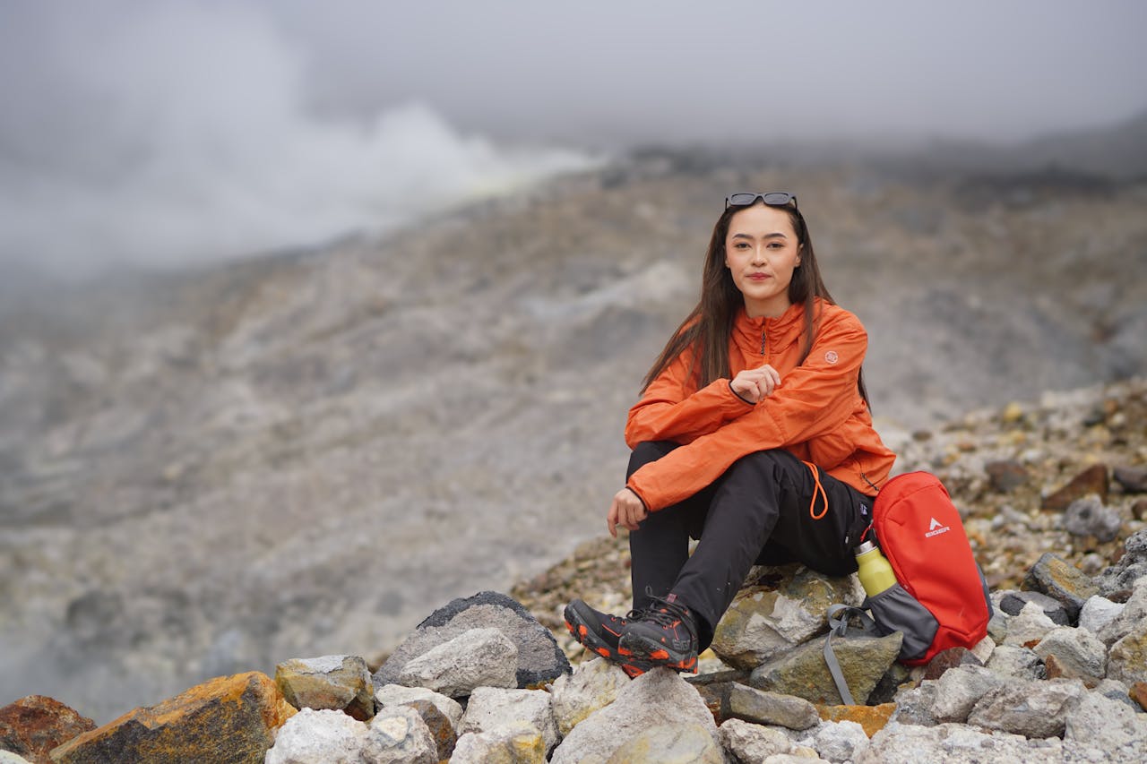 services-03 Woman in orange jacket sitting on rocky terrain, surrounded by foggy mountain landscape.