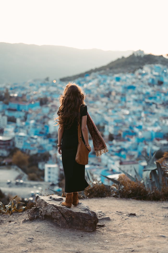 A woman stands on a hill overlooking Chefchaouen's iconic blue buildings at sunset.
