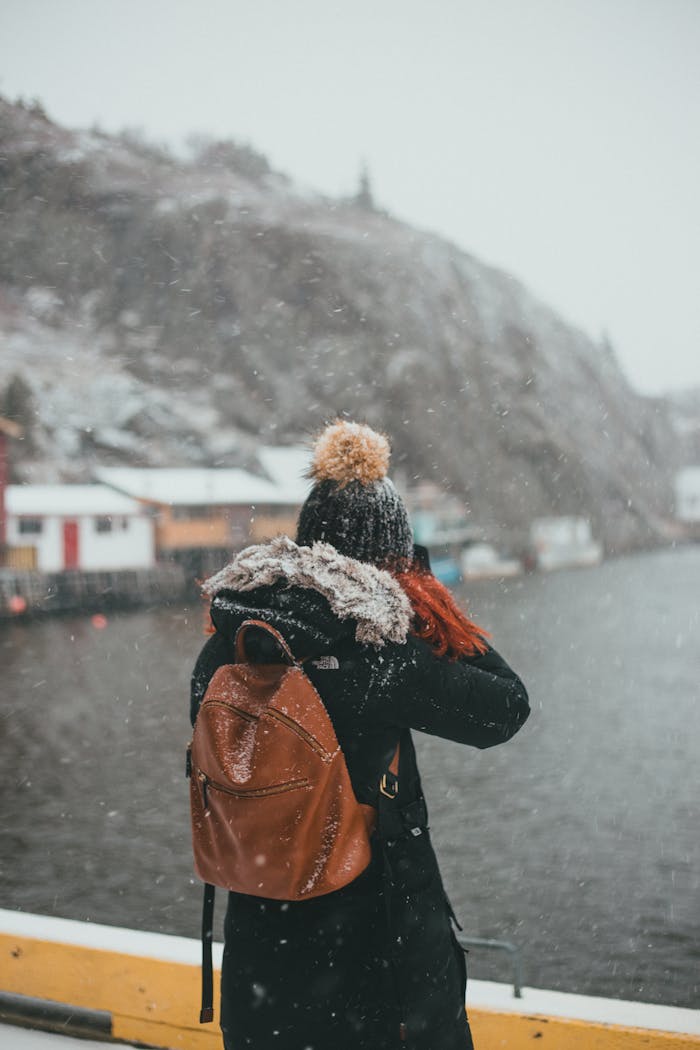 gallery-3 Back view of unrecognizable female traveler in outerwear with backpack standing on embankment and shooting lake surrounded by hills